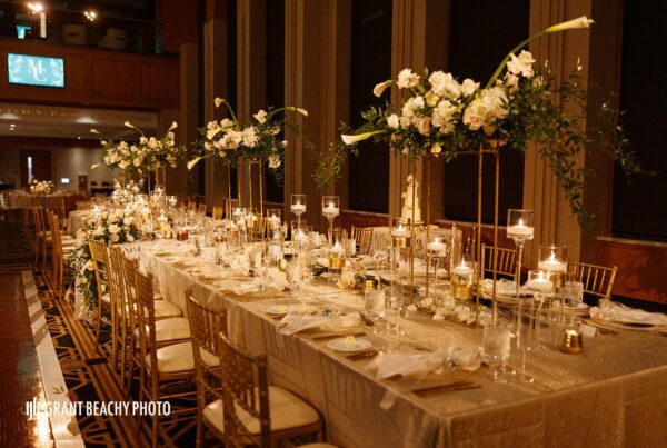 A long shot down the head table with tall floral arrangements filled with white roses, hydrangeas, and long calla Lillie’s and greenery overflowing down toward the table