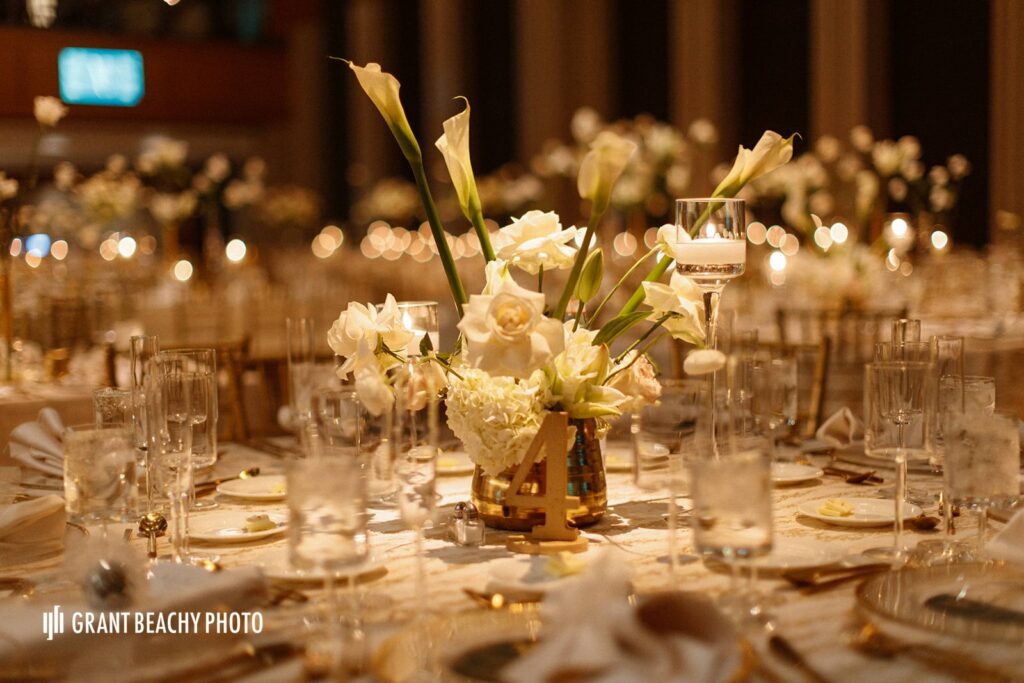 Spotlighted Wedding Tablescape with a White floral centerpiece including Calla Lillies, Roses, and hydrangeas in a Gold Container