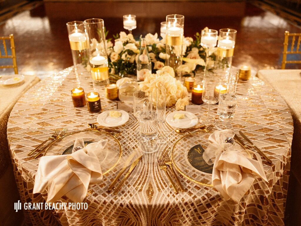 The back side of a wedding sweet heart table: Gold grear gatsby stye linen with glass, gold rimmmed chargers. Framed with floating candles and a white rose and greenery floral design that hangs from the front of the table